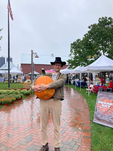 Photo of Tyler standing on a brick sidewark holding a giant pumpkin at the farmers market.
