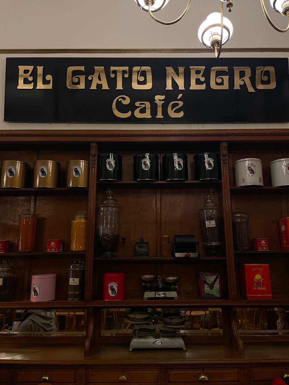 'El Gato Negro' cafe wall full of old colored containers of tea and coffee and spices with a very old scale on display.