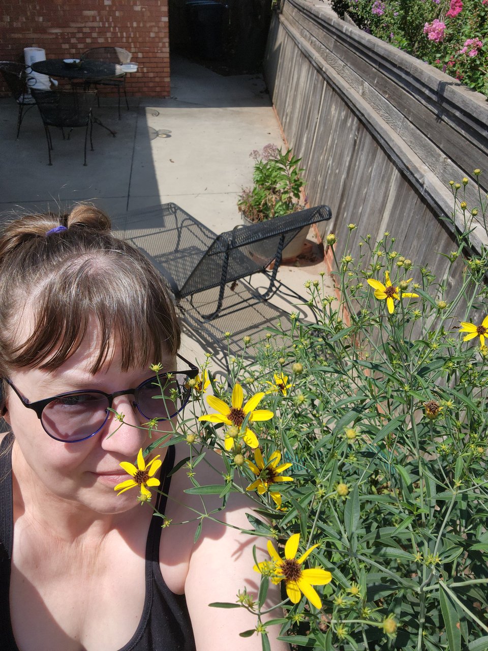 Photo of Sarah in her backyard next to a very tall plant. Sarah is a caucasian person, wearing a black tank top and sunglasses, with her hair in a ponytail. Her plant has small yellow flowers.