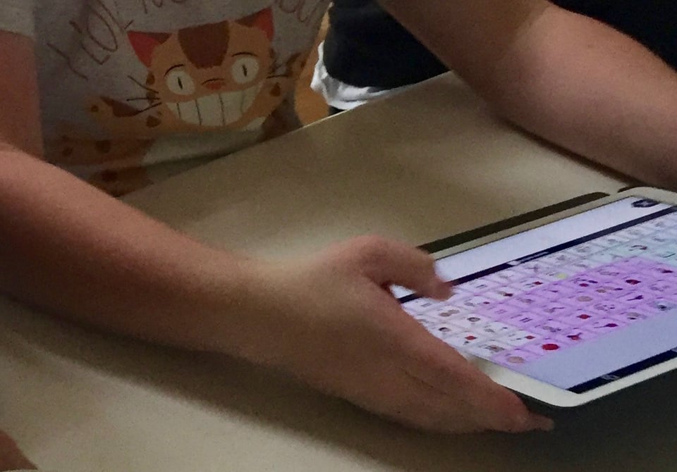 A white teen boy seated at a white counter holds a large iPad displaying the Proloquo AAC app.