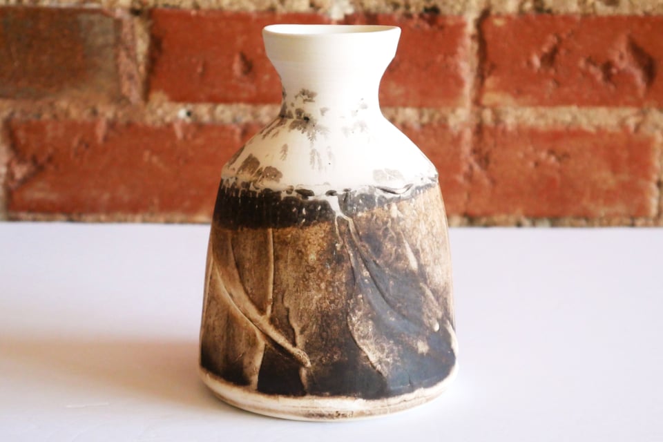 Bottle vase in organic sepia-like matte finish. It has a wide base that rounds back into the foot, holding it gently off the table. The walls are like a smeared-plaster, highlighted by the raku-burned toasty brown splatters. At the shoulder a darker ring,  then white with subtle splashes as the form angles in to a narrow neck with a slightly flared bowl-like lip.