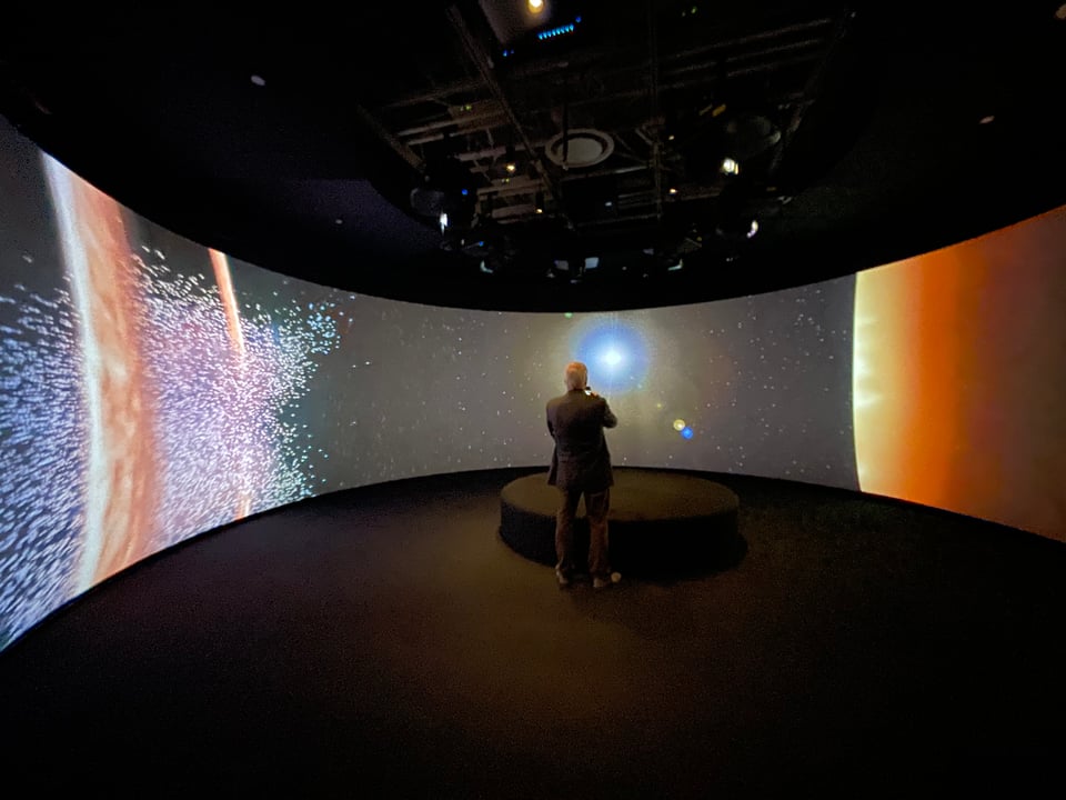 A man stands in a museum gallery surrounded by screens. Multiple images of sci-fi explosions erupt on the screen