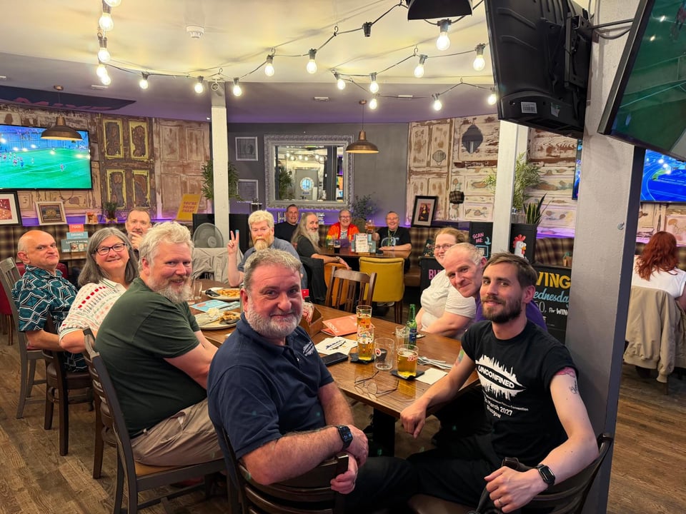 Photo of the people at the Glasgow meetup: a number of people sat at a long table turned to face, and smiling at, the camera.