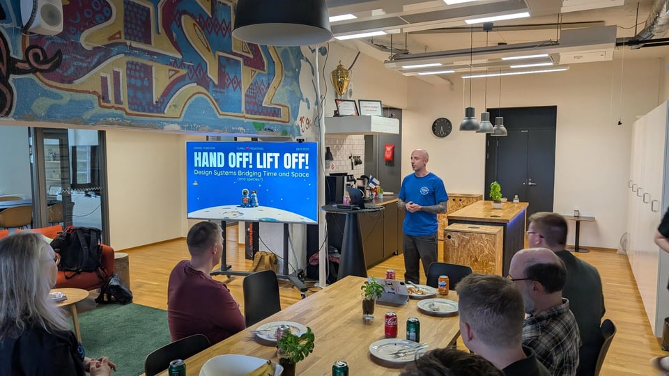 Daniel is speaking in front of an audience in a comfy office kitchen/lounge area. On a screen next to him, a slide has a title of Hand off! Lift off! with a space theme