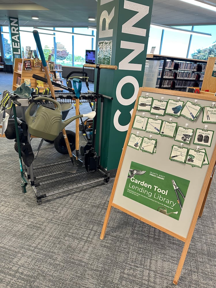 The Garden Tool Lending Library display inside the library. A sandwich board sign hold tags for each of the different tools. Beside it, several tools (e.g. watering can, lawn edger, trowels) are hanging from a metal frame.
