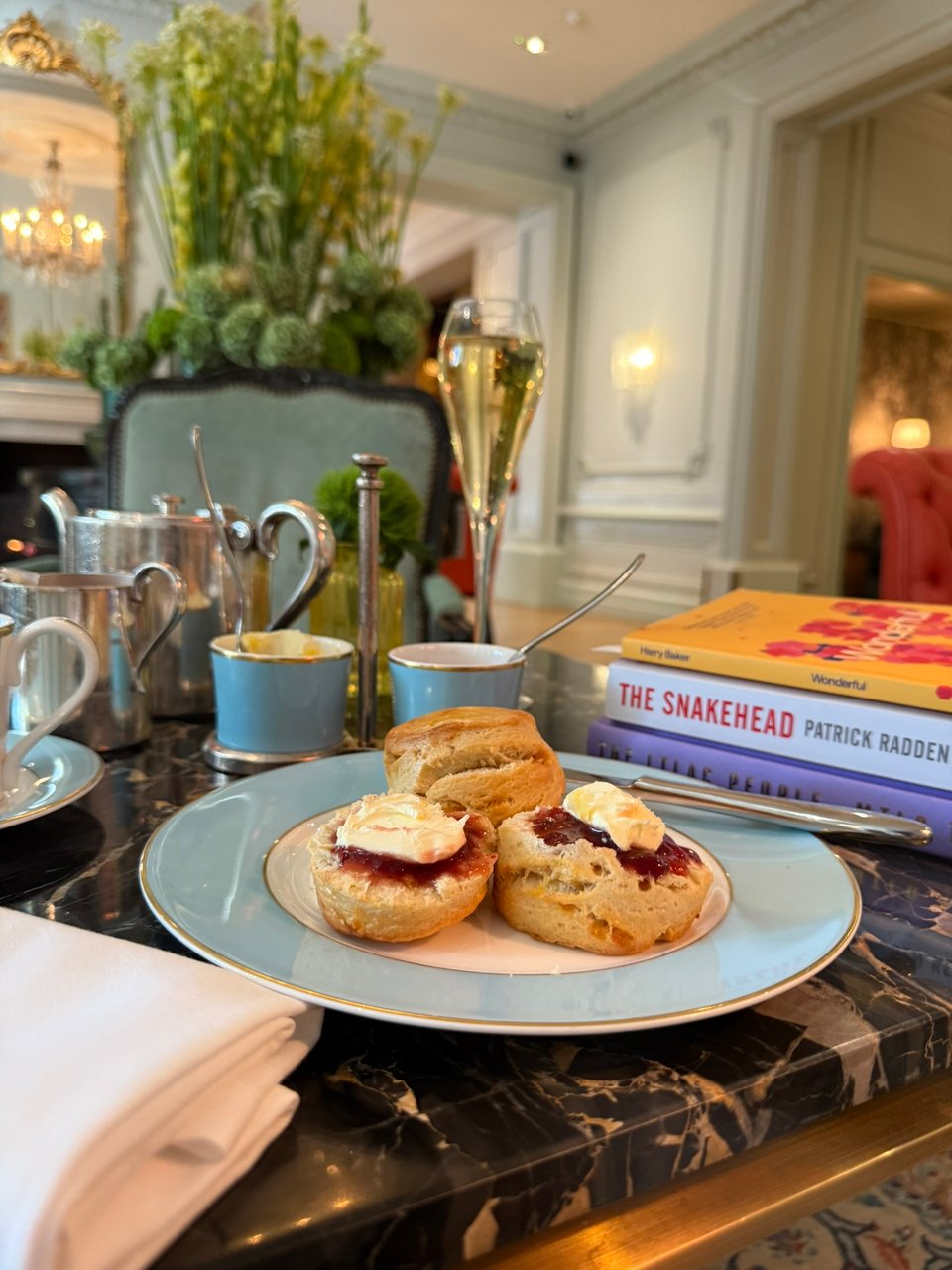 A plate with two scones sits on a table set for afternoon tea. One scone is whole, while the other has been cut open and topped with jam and cream. Also on the table are a champagne glass, a teacup, silver teapots, and a stack of books, including The Snakehead by Patrick Radden Keefe and Wonderful by Harry’s Baker.