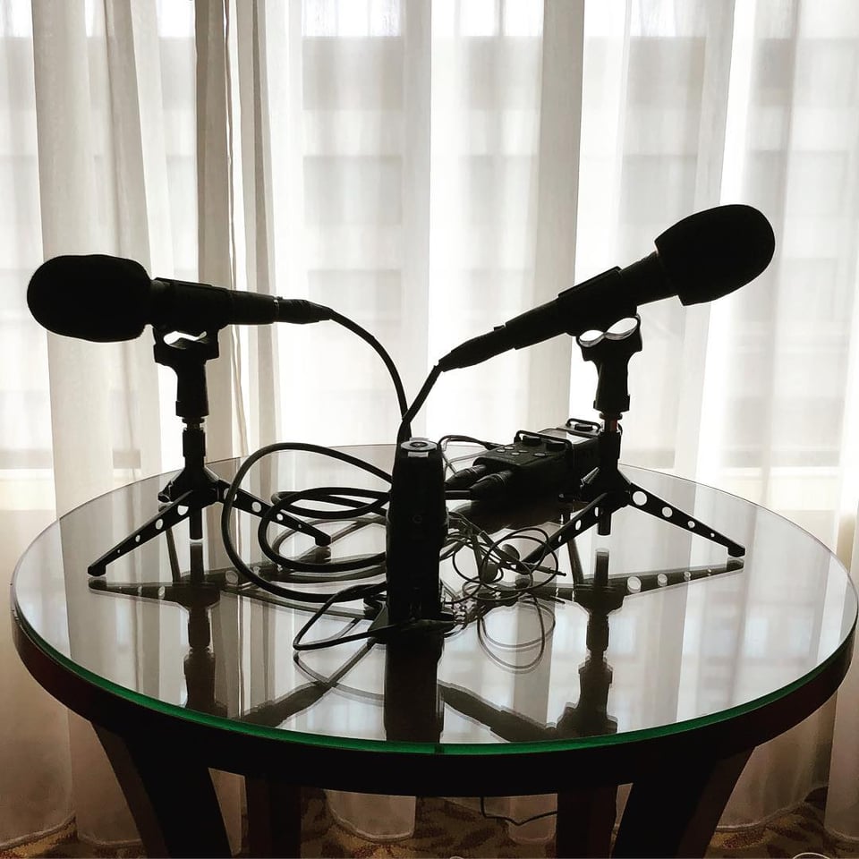 photo of 2 microphones on tripods on a small hotel table. There are two digital recorders on the table, along with various cables