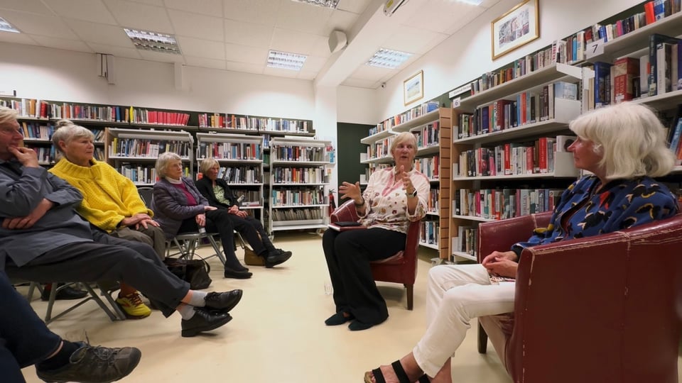 Two women are seated in the front of the room engaged in a conversation, with people listening on in rows of chairs in the audience.