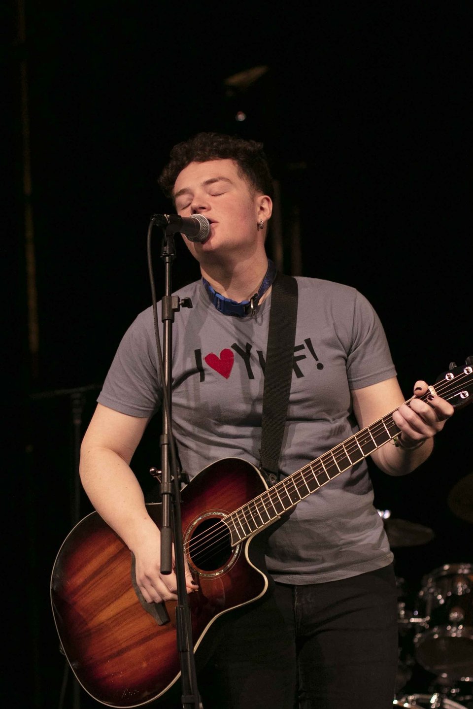 A person with short hair, an earring in their left ear and wearing a gray t-shirt plays an acoustic guitar with a tobacco-sunburst finish while singing into a microphone with their eyes closed.