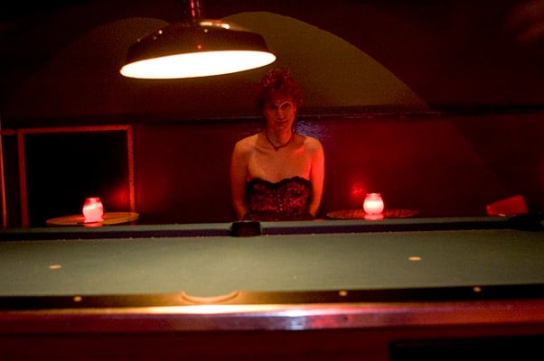 A white trans woman with short curly hair wearing a strapless ballgown and a necklace, with candles on either side of her. She's behind a big pool table.