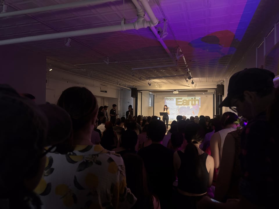 A crowd looks on as Rachel stands at the mic introducing the event. The room has a slight purple glow to it.