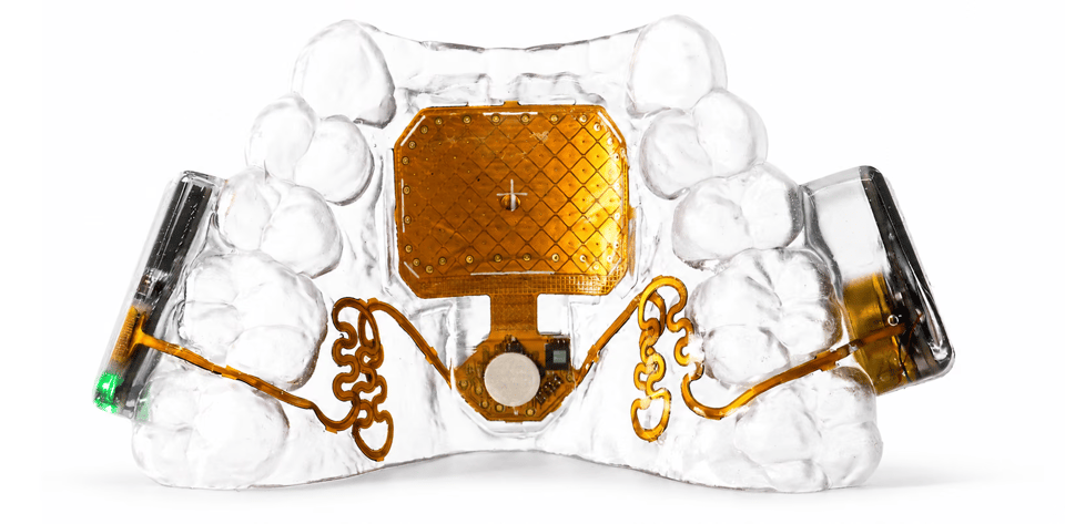 A photo of a transparent mouth guard That is standing upright on its base. We can see a copper square embedded in the roof of the mouth guard, with copper wires snaking out from the copper square to two protrubances on either side of the teeth. It looks very strange and high tech