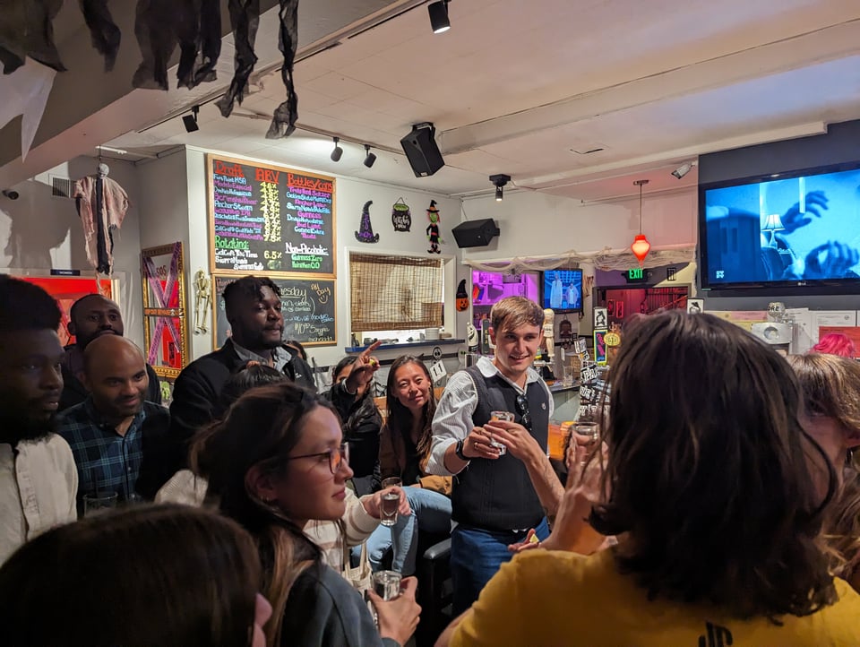 A collection of folks in a bar, some with glasses up in their hands listening to someone speak