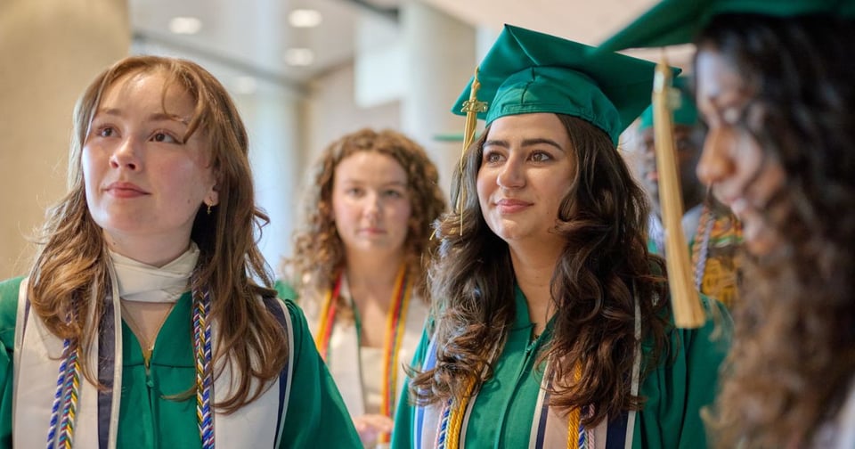 JMC Class of 2026 graduates dressed in green graduation gowns and caps, stand together indoors, likely during a commencement ceremony. Their expressions suggest anticipation or focus as they look upward.