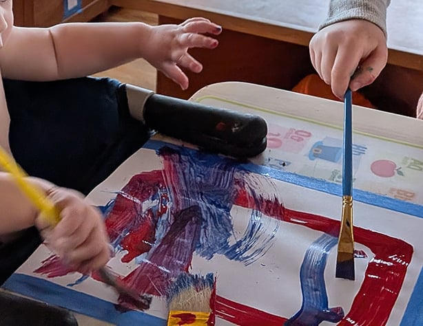 Two young children, only hands and arms visible in this photo, paint together with three different paintbrushes, using reds and blues on a piece of paper taped between them. It's their first collaborative painting and they are having a wonderful time.