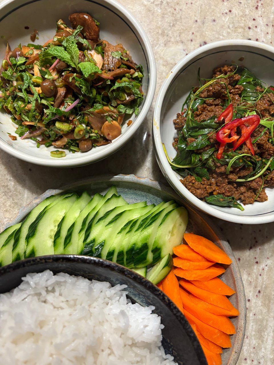 Two bowls, one filled with an assortment of stir-fried mushrooms and herbs and the other with a dry beef curry with fresh peppers and herbs throughout, with a plate of sliced carrots and cucumbers and a bowl of rice.