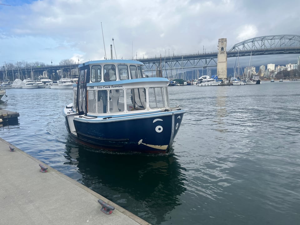 A small blue boat approaches a pier in a crowded harbour. The front of the boat has been painted with eyes and a smile, to resemble a face.