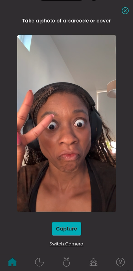 A selfie of me making an exaggerated surprised expression with wide eyes while wearing headphones. I'm holding up a peace sign with one hand. The image is framed within a barcode scanner interface that says 'Take a photo of a barcode or cover' at the top and has a 'Capture' button at the bottom.