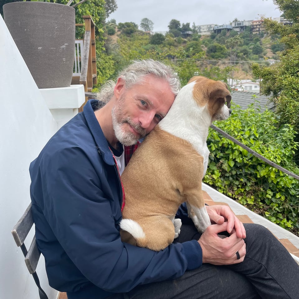 Photo of a man sitting in a chair on a brick patio, with an adorable dog in his lap.