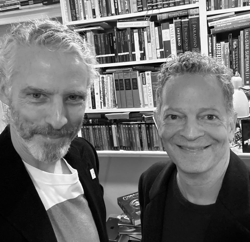 b/w photo of two white men smiling at the camera, with bookshelves behind them