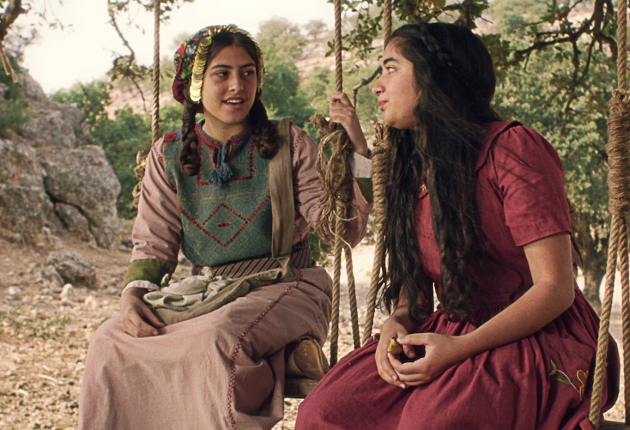 Two young teenage girls in traditional Palestinian dress, sitting on rope swings with trees in the background