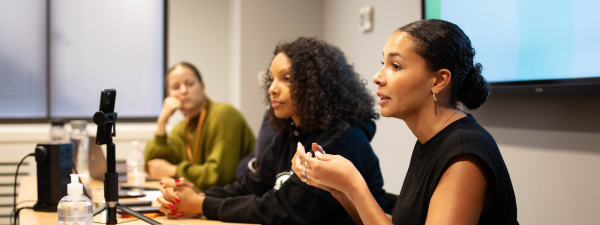 Three woman are at the front of a room, talking. The one furtherest from the camera is out of focus. The other two women are Black and Brown women dress in black. All are sitting down facing to the left