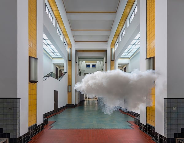 Una nube cruzando un vestíbulo con escaleras a los lados y con algunas columnas y partes del techo con baldosas amarillas. El resto de las paredes son blancas y el suelo es de baldosas rojas con una parte central en color verde aguamarina.