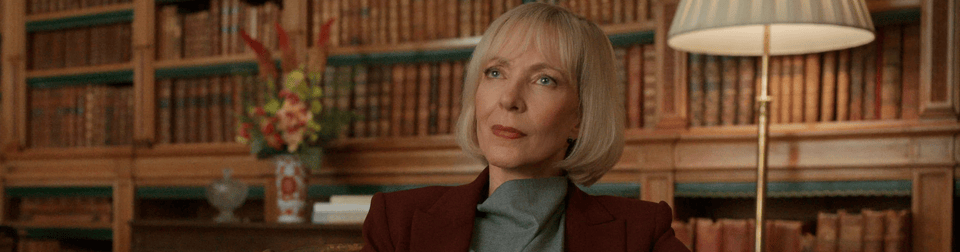 Allison Janney (a white woman with a blonde bob and maroon suit) sits in a brown-toned expensive library with a quietly judgemental look on her face. This is a screenshot from season 2 of the Diplomat.