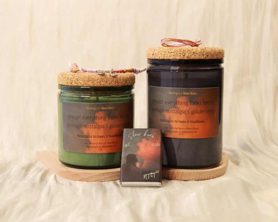 Two candles, one taller than the other, against a cream cloth background. A Slow burn matchbox is leaning against the candles and a friendship bracelet with charms rests on top of the candles. Both have a label with a close-up of rose gold clouds against grey sky and the lyric "almost everything looks better through nostalgia's golden eyes."