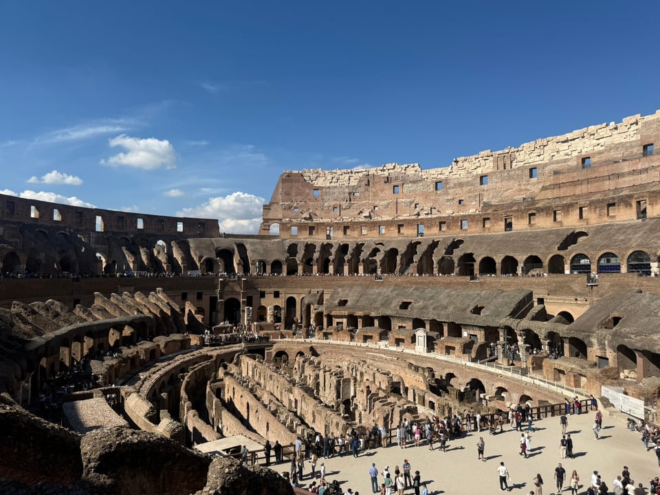 The interior of a large ancient amphitheatre with layered arches and crowds walking along the lower levels.