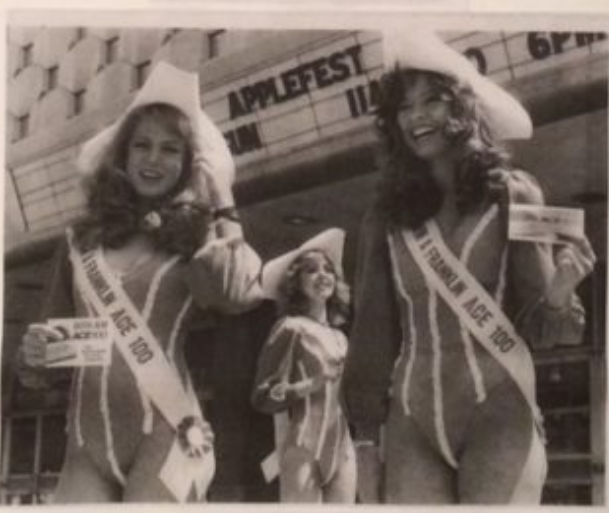 Two female models wearing swimsuits and sahes that say "Franklin ACE 100". They are standing in front of a convention center and the marquee says "Applefest" in the background.