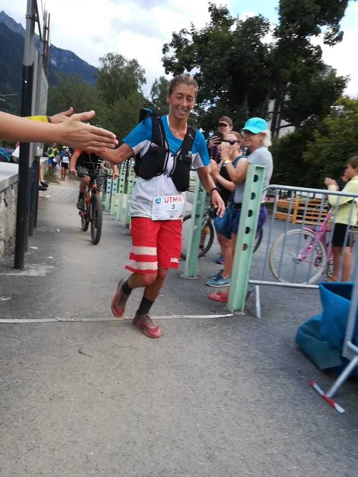 A woman running towards the camera, passing what appears to be the finishing line of a race. She is wearing red shorts, red shoes, a blue and white jersey, and is reaching out to give a high-five to a hand extending in the left frame of the photo. She seems tired by happy