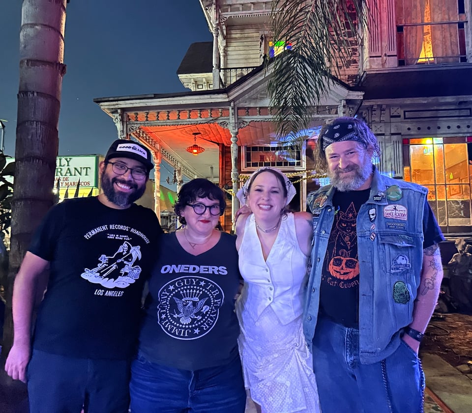 standing in front of the haunted house are rudy, a mexican man in glasses with a beard and a big smile; annika, a white woman in glasses with short dark hair; nikki, a white woman wearing white and a radiant smile; and will, a white man in a denim vest and bandanna with a grizzly beard, looking like he might work the line in the kitchen.