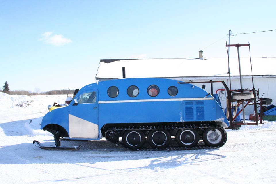 bright and sunny photo of a bright blue vehicle that is hard to describe; a trucklike cab poised on a pair of rugged black skis, curving into a strange cabin with porthole-like windows over a heavy-duty (yet somehow still cute) tread