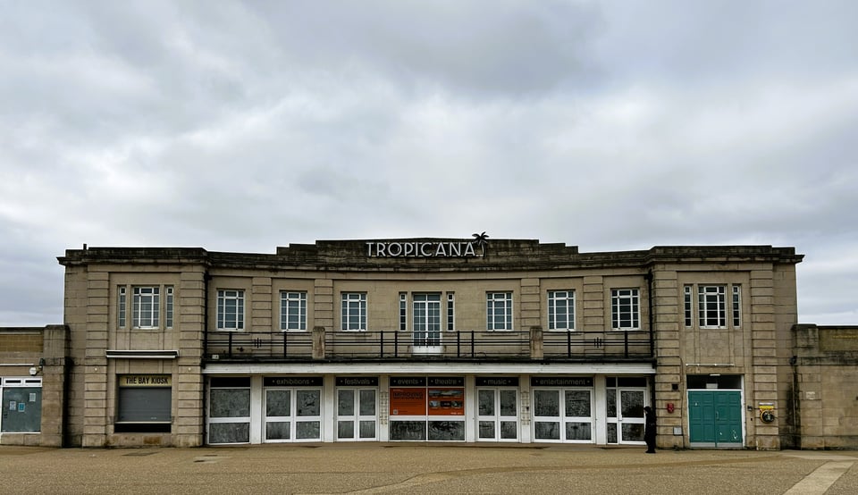 A wide low two storey building facade. The ground floor is mostly obscured glass doors. Above is a slightly set back curved balcony with a stepped parapet holding the name 'tropicana'.