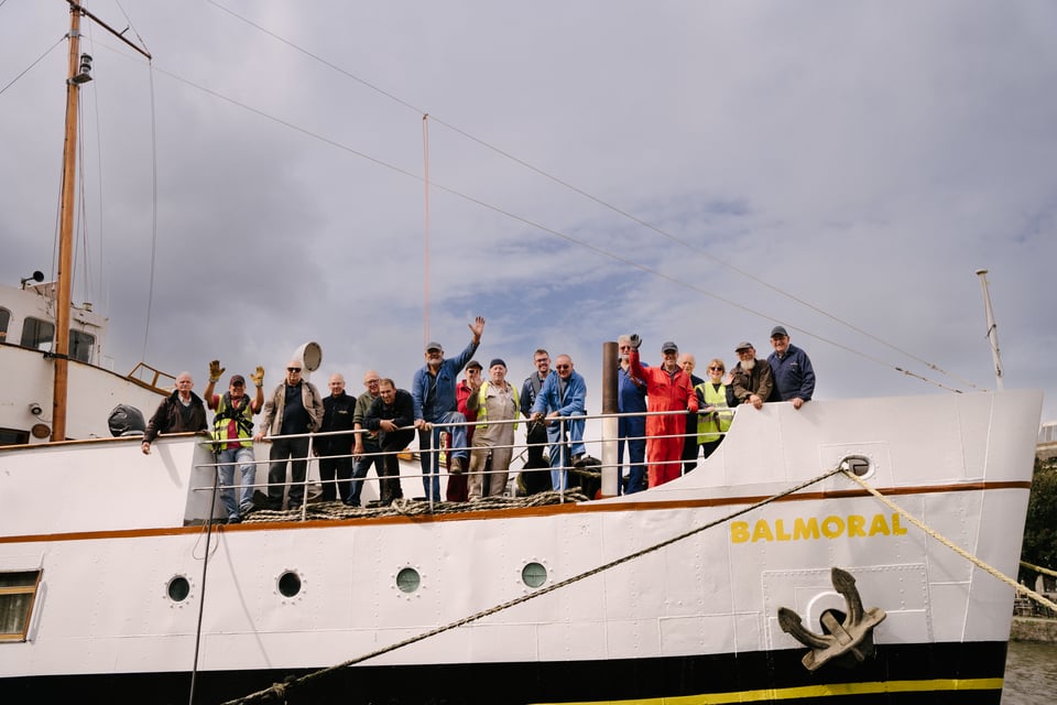 A group of volunteers stand on the bow of a ship. We see the sign 'Balmoral' to the lower right of the image and a cloudy sky in the background. Some of the volunteers are waving at the camera.