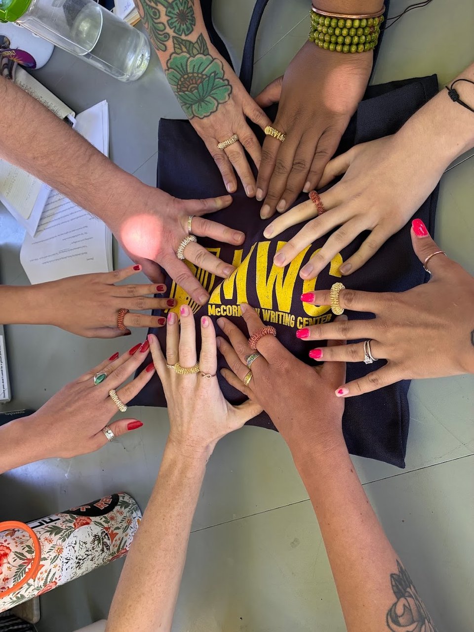 An image of nine hands and arms meeting each other, each hand and arm different, but all with a little fidget spiral thing on one finger