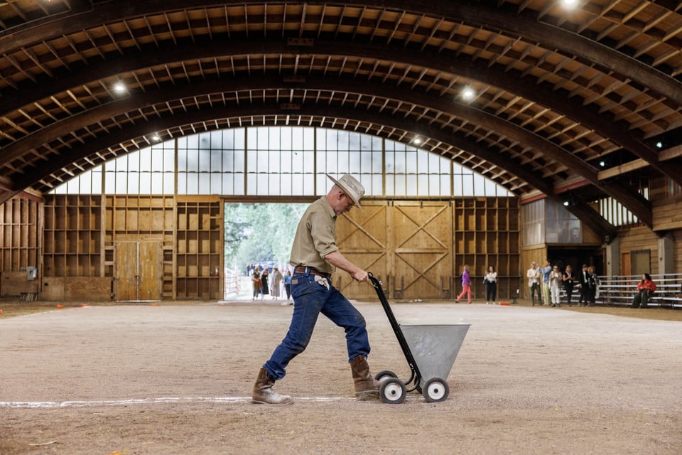 Performance artist Matthew Barney is in a domed, wooden drill hall-turned-barn on a Colorado ranch, centered in the image with an open entrance at the back. He is wearing Western/ranching attire (jeans, boots, cowboy hat) and pushing an old fashioned line-marker machine (for sports playing fields), leaving a white line in his wake. There are bleachers on the right side of the space. Trees and a small group of performers are visible through the entrance at the back.