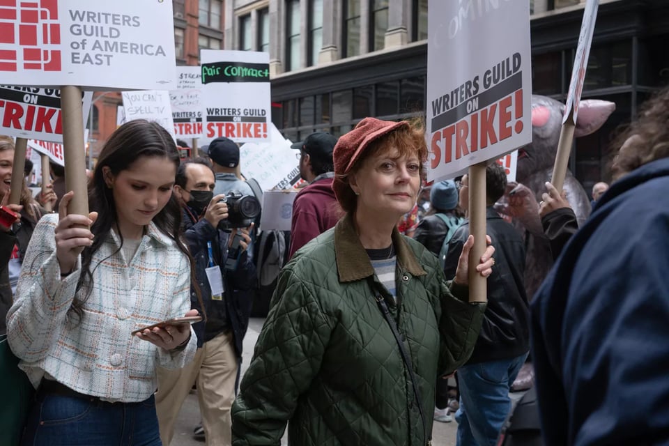 Susan Sarandon walks among many other people at a march, holding a sign that says "Writers Guild On Strike"