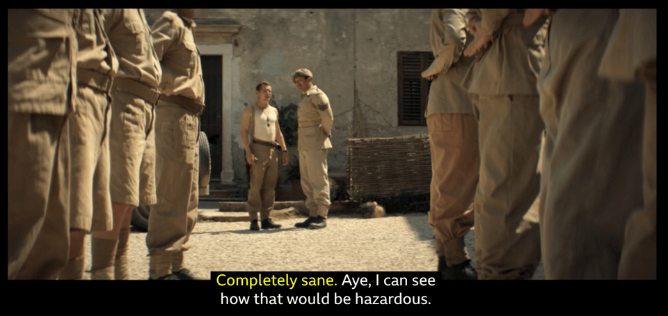 A screenshot from the TV show SAS Rogue Heroes. In the foreground are two rows of soldiers standing to attention. They are out of focus but we can see their upper legs and lower torsos. The camera is focused on two soldiers having a conversation in the background. These conversing soldiers are framed by the two rows of soldiers in the foreground. We also see some subtitle text, which says, "Completely sane." "Aye, I can see how that would be hazardous."