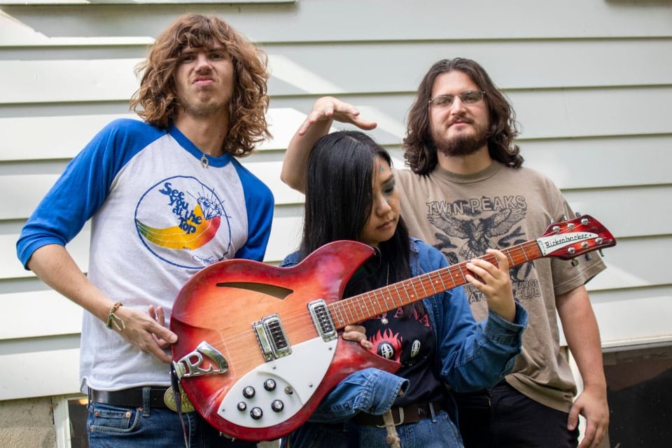 A woman with long dark hair looks at her fingers on the fretboard of a Rickenbacker guitar while a man to her left with glasses and a beard holds his open palm just above her head. To her right, a man with shaggy hair wearing a white t-shirt with blue three-quarter length sleeves has his right hand on the guitar.