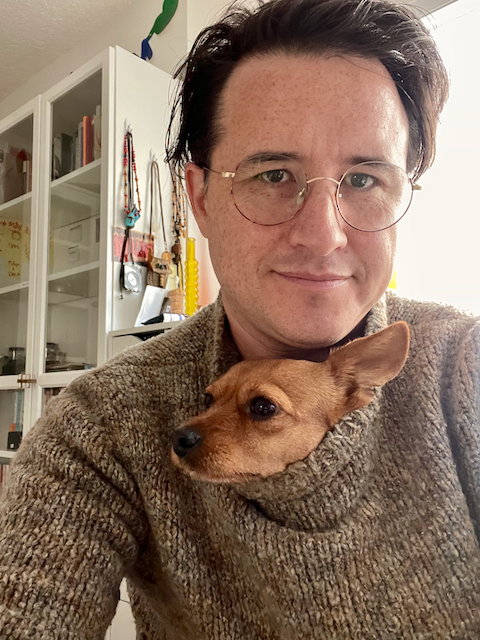 Me, a freckled nonbinary person with dark, ear-length hair and round glasses, framed from head to mid torso, wearing a light earth-toned mock-turtleneck sweater. My little dog, a chihuahua-dachsund mix with coppery coloring, also has her head coming out of my sweater's neck hole under my chin. Her one visible ear is almost as big as her whole head.