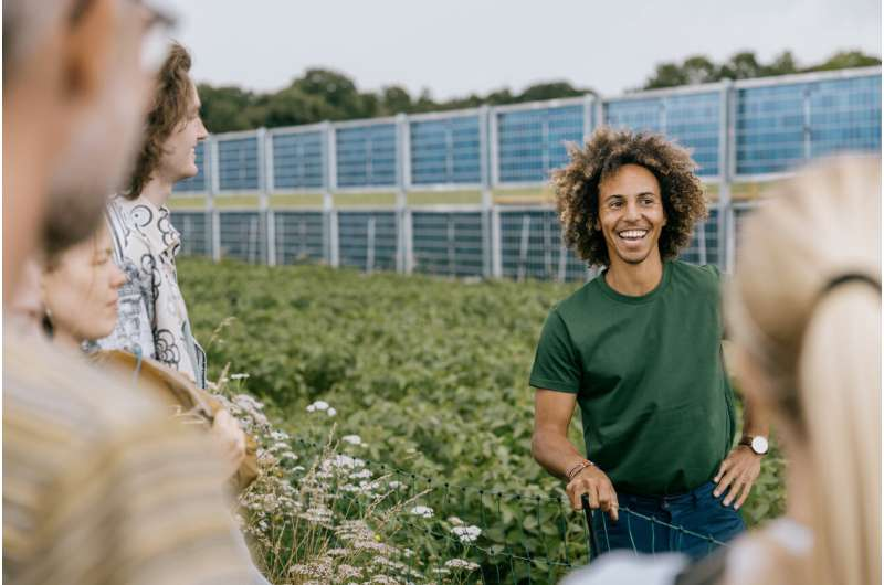 A smilling man in a green tshirt stands in a green field, he is facing the camera and talking to several people, we can see the backs of their heads because they are facing him. In the far background is a row of solar panels that stand up vertical, like a fence