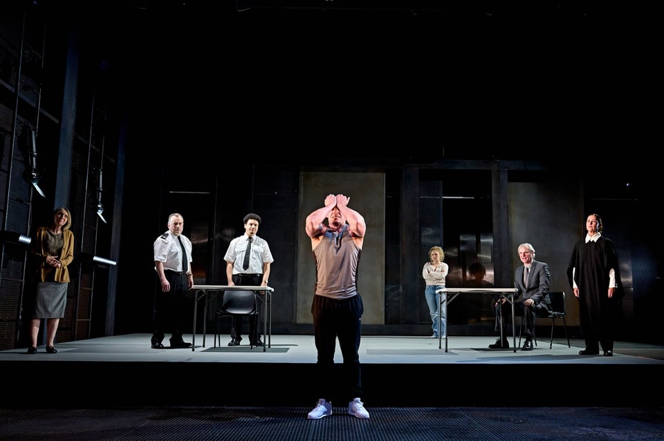 Samuel Edward-Cook, as Raoul Moat, stands on a grate in front of the stage, clasping his forehead. He's wearing a grey gym singlet, black trackpants and white sneakers. He's very buff. Behind him, on a grey, shadowy stage, six people are standing, three on either side. From left to right: an older woman wearing a brown cardigan and grey dress; two police officers wearing white shirts and black ties; a woman leaning against a wall with her arms crossed, with blonde curly hair in a ponytail, wearing a cream cardigan and blue jeans; an older man in a suit, sitting down at a trestle table; and a woman dressed in barrister's robes standing next to the older man.