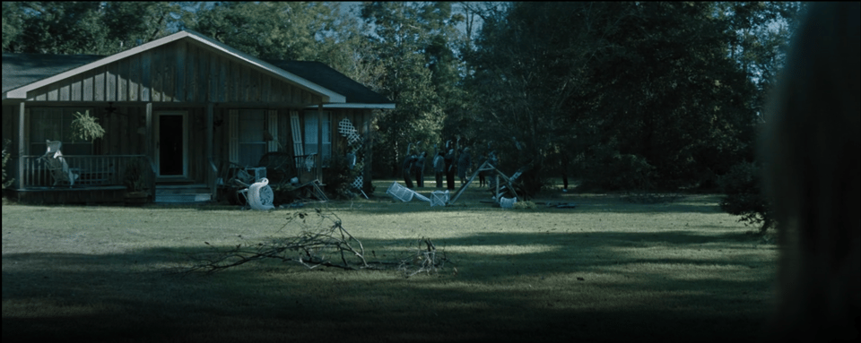 Most of the frame is filled with Brynn's neighbor's house, many of its contents spilling haphazardly across the yard. A group of people from town stand in the side yard, their right hands raised palm up to the sky and their backs arched backwards.