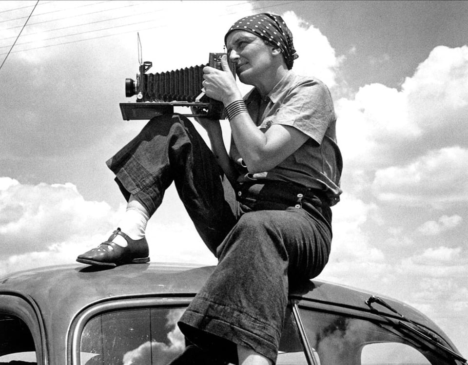 A photo of the photographer Dorothea Lange with her field camera as she sits on top of the roof of a car.