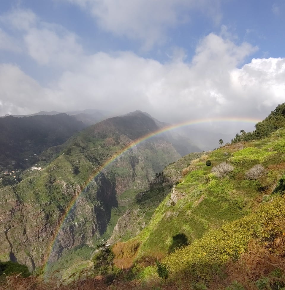 Blick auf steil bergige Landschaft, darüber locker bewölkter Himmel. Vom Tal her zieht sich ein Regenbogen an den Hang.