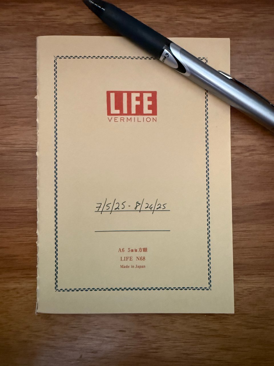 photo of a LIFE Vermilion A6 notepad with a pen lying across it to keep it shut. It has the dates 7/5/25-8/26/25 written on the cover