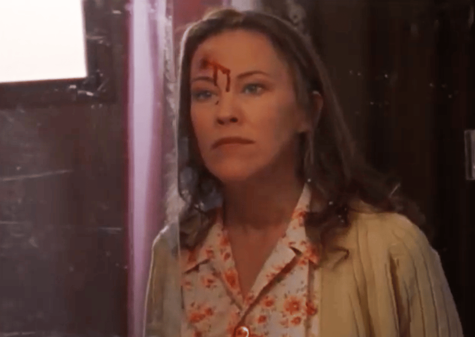 Catherine O'Hara stands in front of her bathroom mirror in a floral blouse and beige cardigan, looking at the sizeable bullet wound in her forehead and the blood trickling down to her nose.