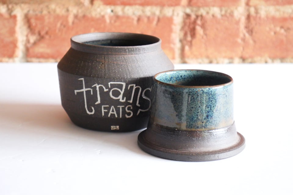 An open butter bell in two pieces, the upright jar and overturned lid revealing a bell inside – now upright. On the side of the jar it says trans fats in a hand-carved slab serif inlayed withc white. The outside surfaces are a rich brown raw clay with subtle texture, while the insides are glazed in organic floating blues.
